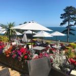 Sunny seaside terrace with colourful flowers, parasols, and ocean view in the background