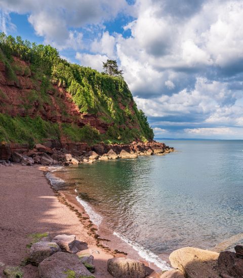 Scenic view of a tranquil beach cove with red cliffs and lush greenery under a partly cloudy sky