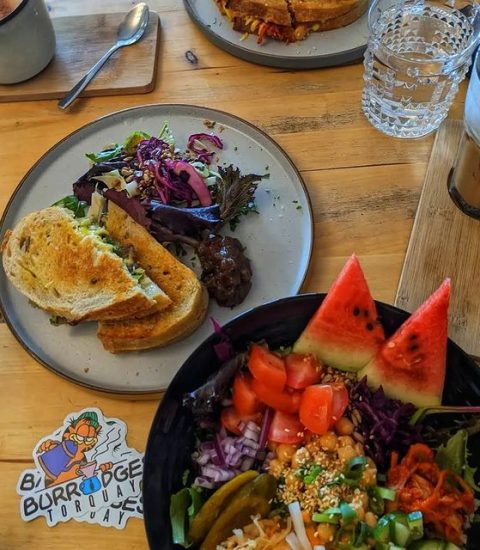 Colourful salad bowl and sandwich with mixed greens on a wooden table, accompanied by drinks and cutlery