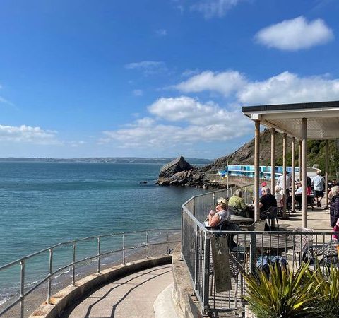 Coastal view with people enjoying a seaside cafe on a sunny day, rocky outcrops in the distance under a blue sky