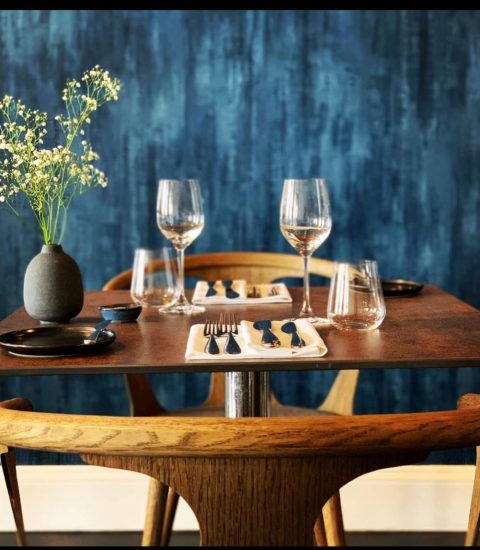Elegant table setting with wooden chairs, wine glasses, cutlery, and a vase with flowers against a blue textured wall