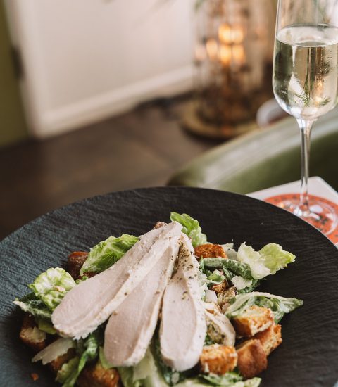Chicken Caesar salad with croutons and Parmesan served in a black bowl, accompanied by a glass of sparkling wine