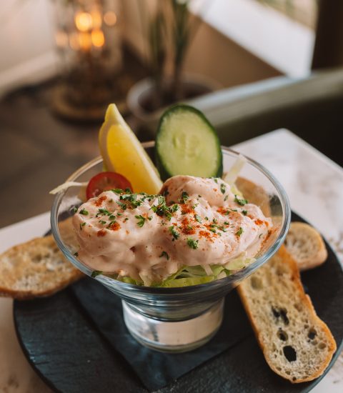 Prawn cocktail with lettuce, lemon wedge, cucumber slice, and crostini on a marble table in a glass dish
