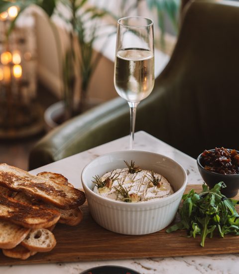Cheese board with artisan bread, baked camembert, chutney, rocket and a glass of white wine on a marble table