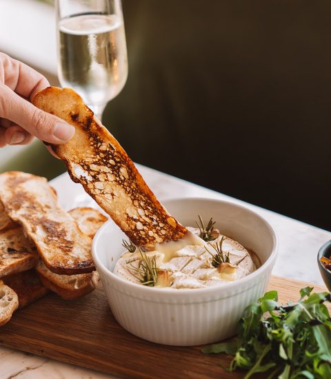 Hand-dipping toasted bread into baked camembert with herbs, alongside chutney and fresh salad on a wooden board