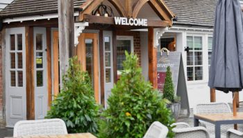Entrance of a welcoming café with outdoor seating, plants, and signage against a traditional brick wall backdrop