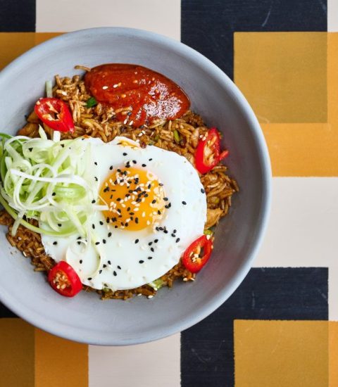 Bowl of fried rice topped with fried egg, sliced cucumber, red chillies, and sauce on patterned background