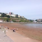 A scenic view of Torquay beach with people relaxing on the sand, the hillside, and a Ferris wheel in the background