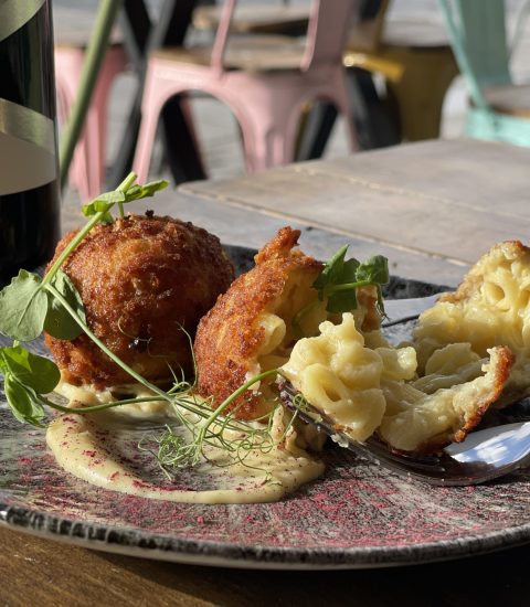 Gourmet macaroni cheese bites with creamy dip and herbs, paired with a bottle of Merlot on a rustic table setting