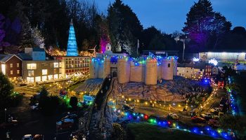 Miniature festive village with castle, colourful lights, tiny figures and shops at night, surrounded by trees