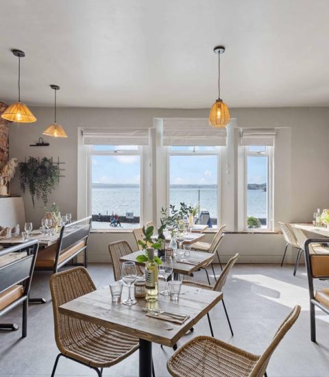 Cosy coastal restaurant interior with ocean views, rustic decor, wicker chairs, and warm lighting ambiance