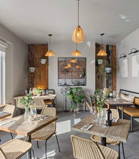 Cosy restaurant interior with wooden tables, wicker chairs, hanging lights, and potted plants by large windows