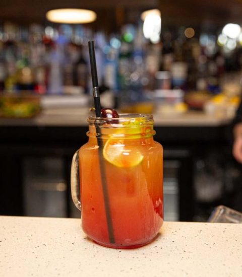 Colourful cocktail in a mason jar with fruit garnish on a bar counter, blurred bartender in the background