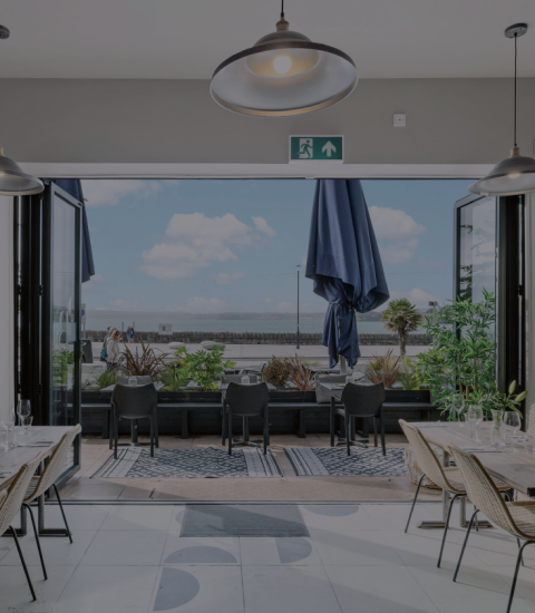 Modern seaside restaurant interior with open doors to a patio, natural light, and stylish decor