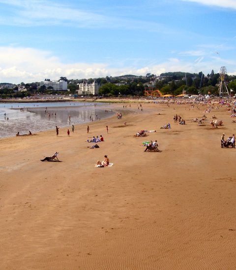 Sandy beach on a sunny day with people relaxing, calm sea, and amusements in the distance, Torquay, Devon