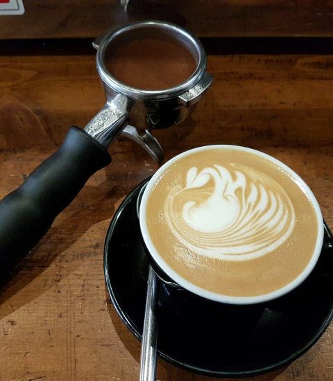 Latte art with a swan design in a black cup on wooden table beside espresso portafilter