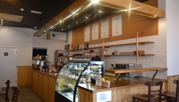 Cosy modern café interior with wooden counters and seating, two staff members behind the counter preparing orders