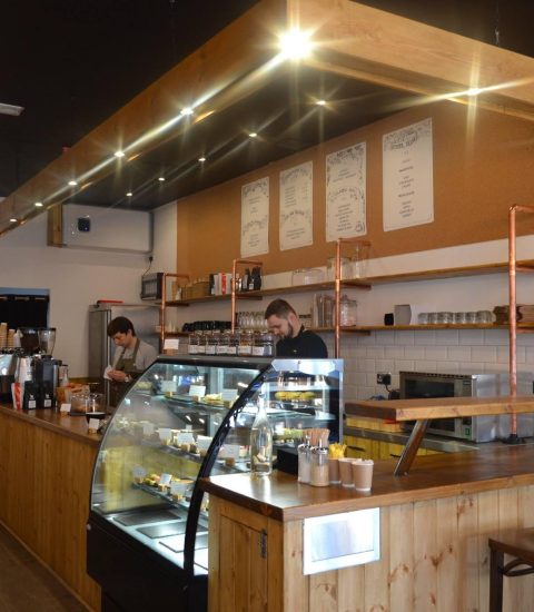 Cosy modern café interior with wooden counters and seating, two staff members behind the counter preparing orders
