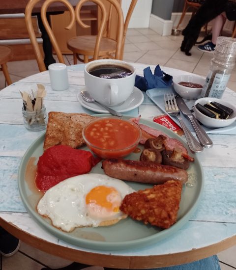 Full English breakfast with fried egg, sausage, bacon, beans, mushrooms, hash brown, and coffee in a café setting