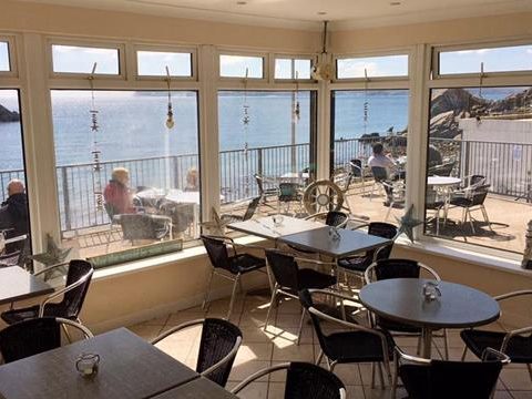 Seaside café interior with large windows overlooking outdoor seating and ocean views, patrons enjoying the sunny day