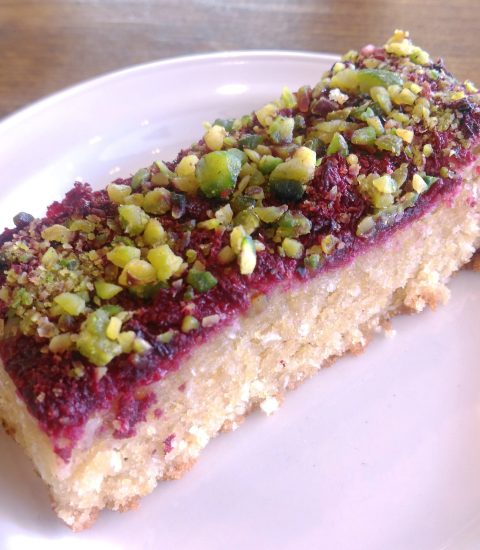 Slice of raspberry and pistachio cake on a white plate, highlighting vibrant colours and crumbly texture