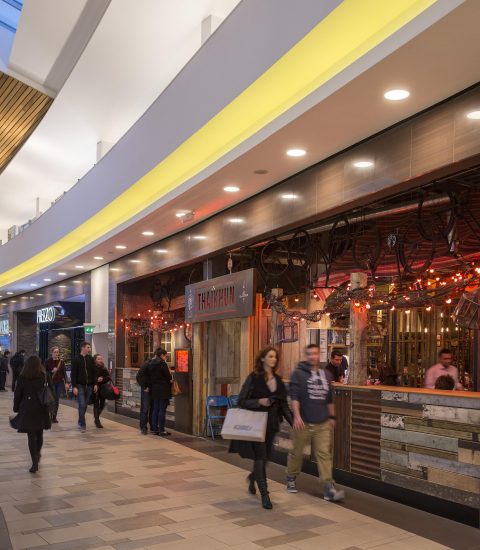 Shopping centre with people walking by a Thai restaurant featuring bright lights and a rustic wooden exterior