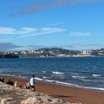 Beachgoers enjoy a sunny day at Torquay beach, with coastal town in the background and blue skies above