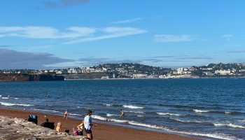 Beachgoers enjoy a sunny day at Torquay beach, with coastal town in the background and blue skies above