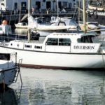 Boats moored at a vibrant marina, reflecting on calm water, with buildings and a ship in the background