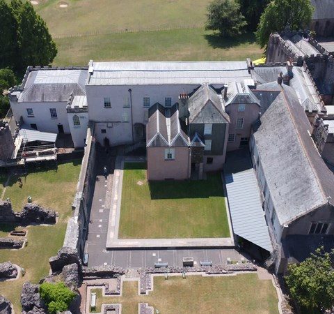 Aerial view of a historic building with green lawns, surrounded by ancient stone ruins and lush trees
